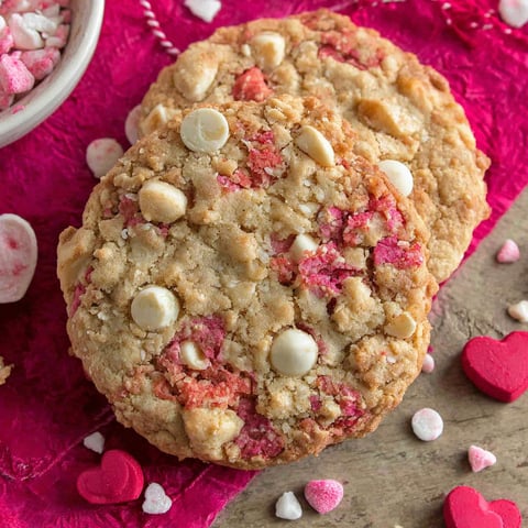 Two cookies with white chocolate chips and red icing on a pink surface.
